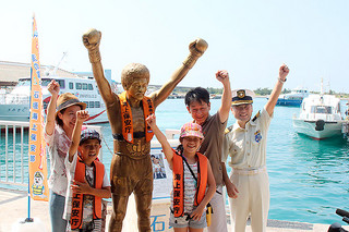 救命胴衣を着せられた具志堅用高の銅像と記念撮影をする観光客ら＝16日午前、同離島ターミナル