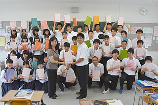 学校全体で勉強を頑張る空気をつくろうと真久田絹代校長（中央左）が直接手渡している自学自習ノートを手に、笑顔の八重高１年２組の生徒たち＝25日午前、同校