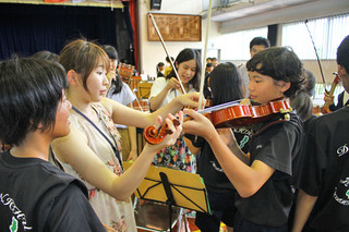 東北ユースの楽団員（左から２人）からバイオリンの手ほどきを受けるマーチングバンド部の部員＝９日午前、県立石垣青少年の家体育館