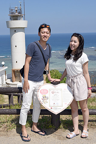 昨年７月の県内初認定を受けて設置された「恋する灯台」の看板と記念写真に納まる観光客。後ろは平久保崎灯台＝19日午前、同灯台